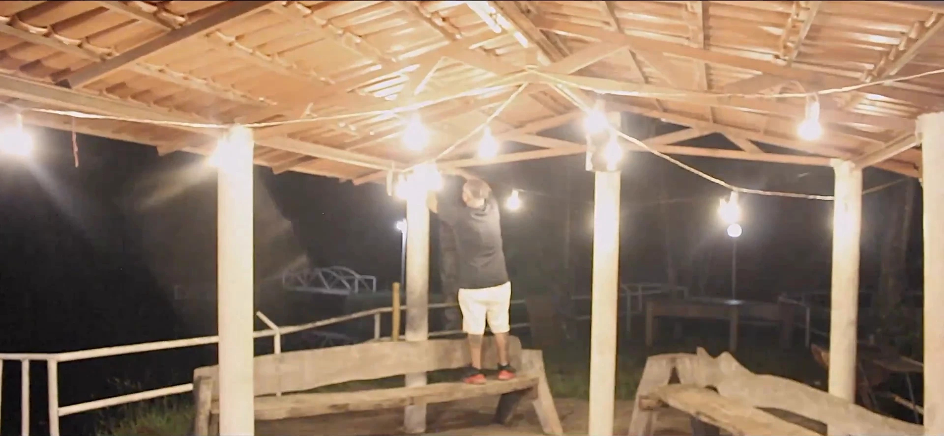 Pavilhão de madeira à noite, com lâmpadas de filamento no teto e uma pessoa ao fundo. O plano de situação mostra o lugar antes de a festa começar.