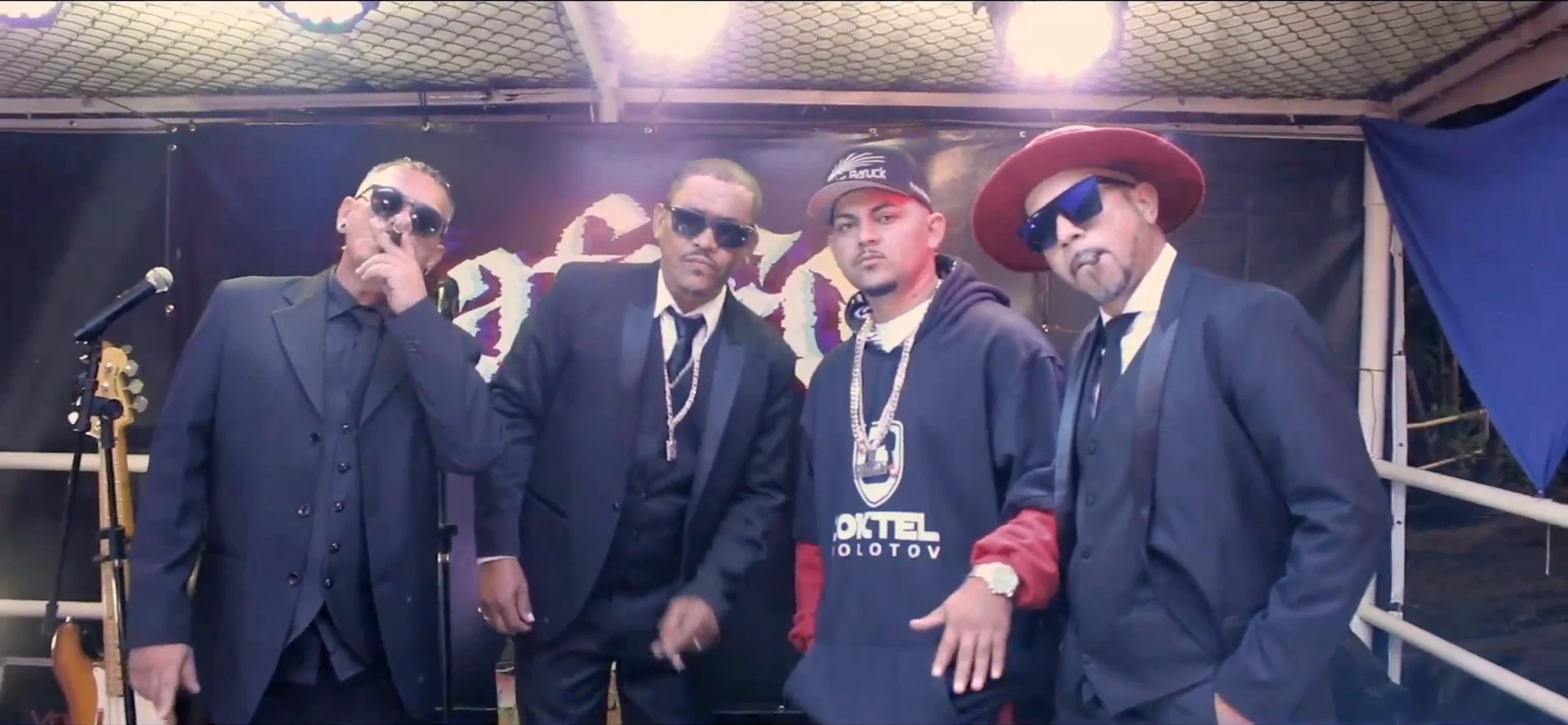 Quatro homens posando no palco, três de terno escuro e um de moletom, com correntes e óculos escuros. Ao fundo, o letreiro gótico "Cafofo" sob luz violeta.