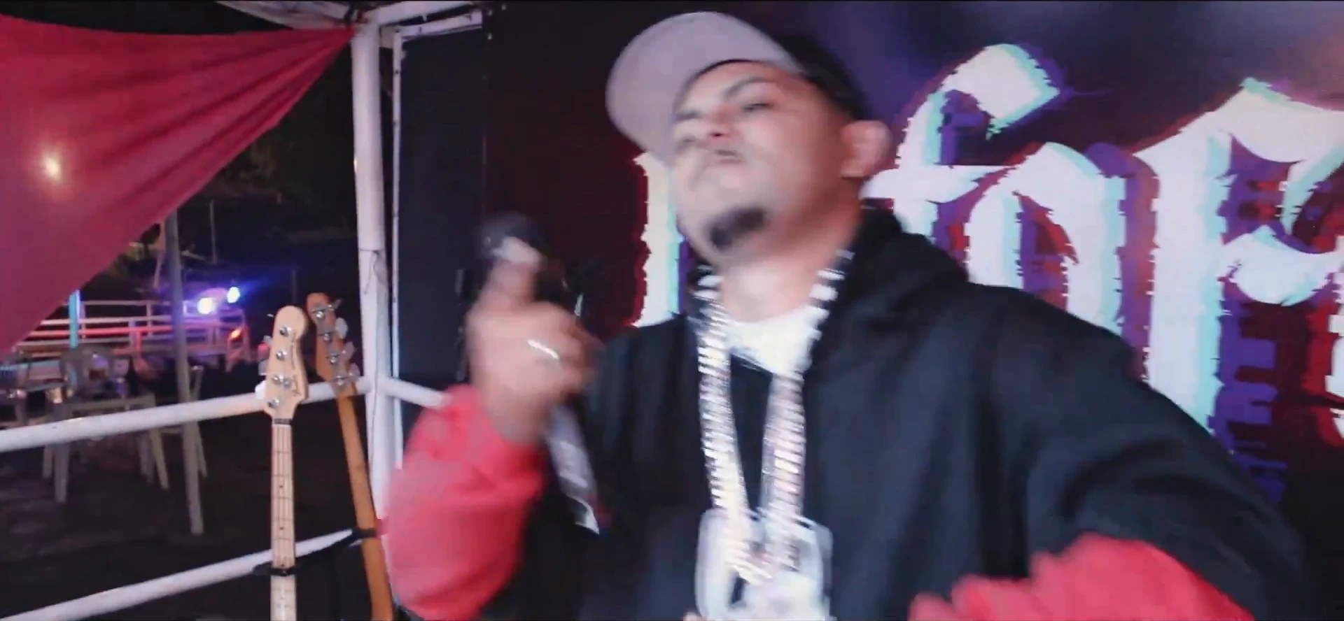 MC de boné e correntes grossas canta ao microfone, em movimento, diante do letreiro gótico. Ao lado, dois baixos apoiados no guarda-corpo e um pano vermelho.